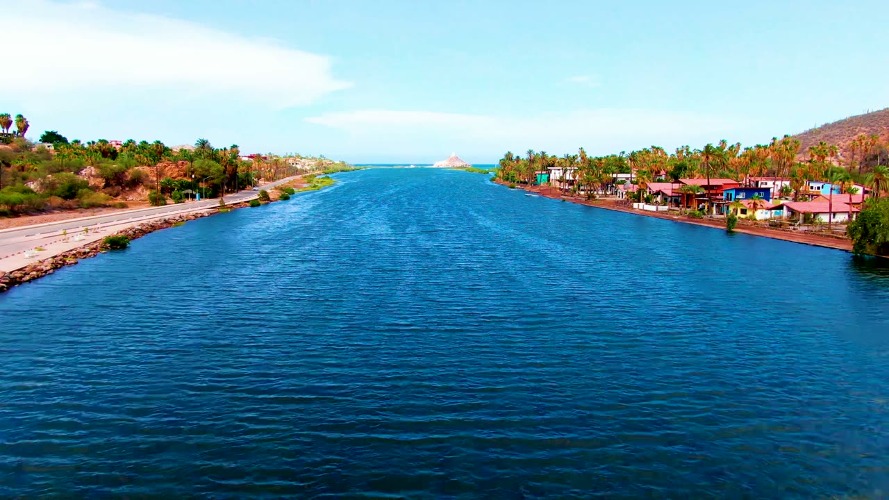 Opening aerial frame over a blue canal in Mulege, Baja California Sur, with a road along the left bank, colorful low buildings and palms on the right, dry hills, ocean on the horizon, and no visible people.