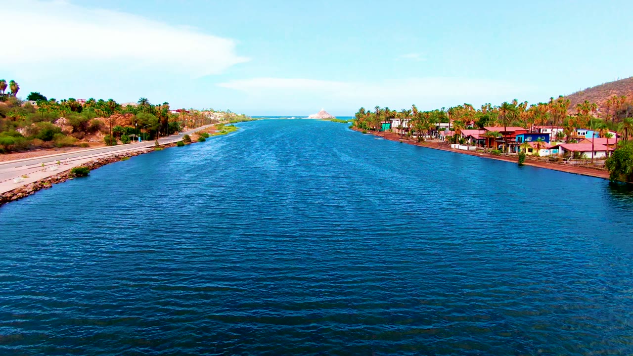 Wide aerial canal view with rippled blue water, road, buildings, palms, and hills.
