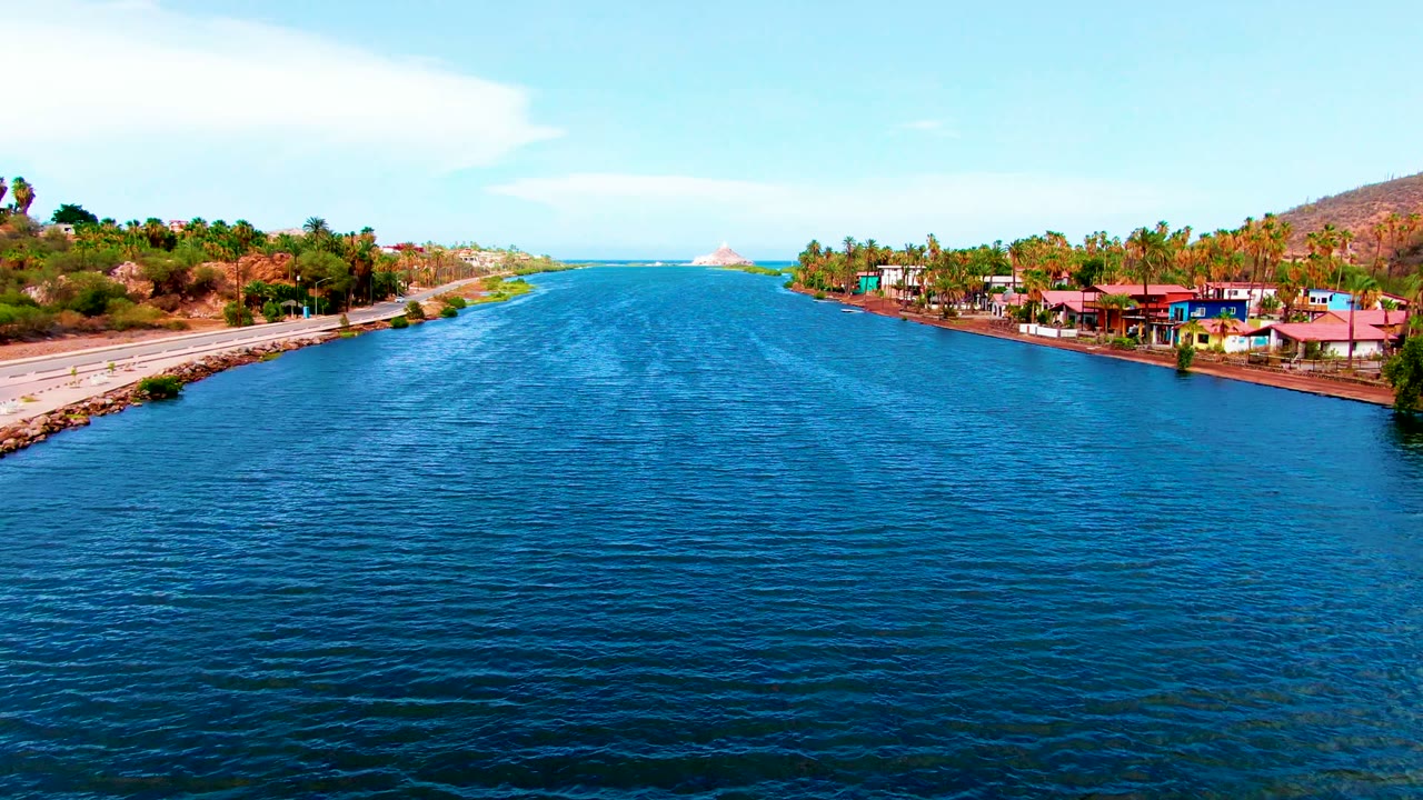 Aerial frame over the Mulege canal with textured blue water in the foreground, a paved road and rocky bank at left, palm-lined buildings at right, low dry hills, ocean in the distance, and no visible people.