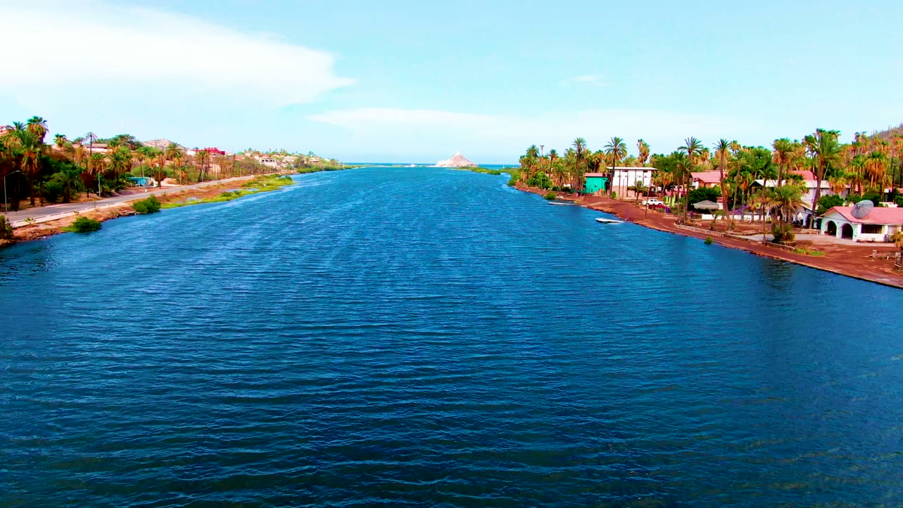 Wide aerial frame over a blue canal in Mulege, Baja California Sur, with palm trees, shoreline houses, a road edge, distant coast, and no visible people.