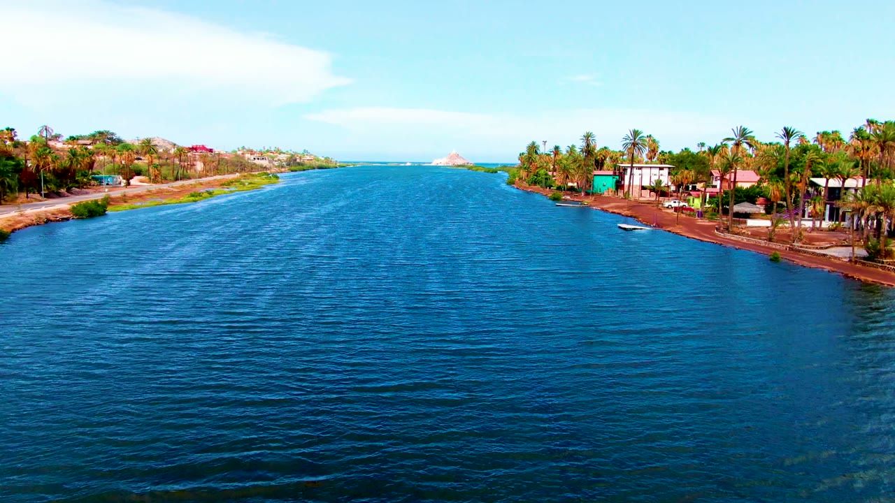 Elevated Mulege canal frame with open blue water in the foreground, palm clusters and houses along the right bank, a lower left shoreline, and no visible people.