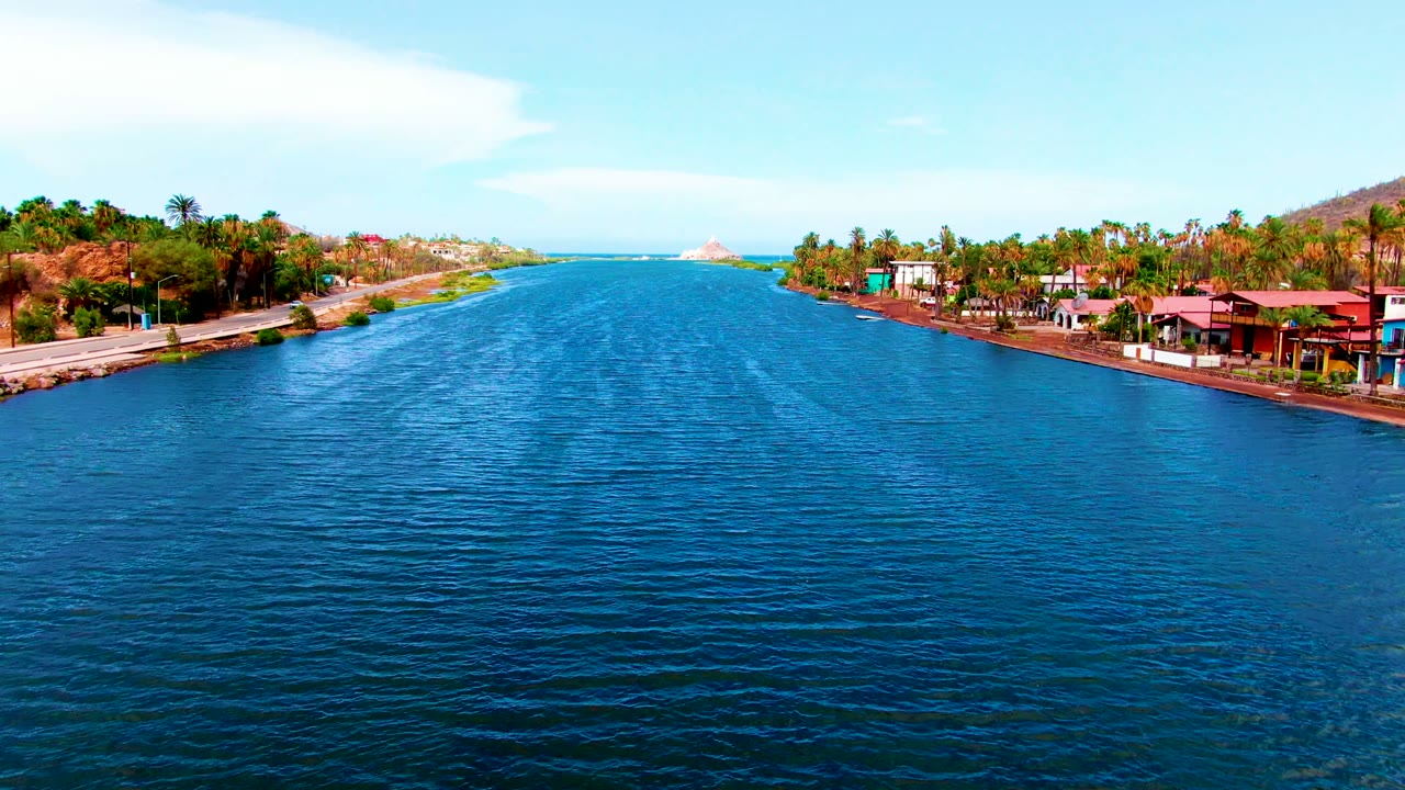 Opening frame of a Mulege, Baja California Sur canal with rippled blue water centered between palm-lined banks, low waterfront buildings, distant hills, and no visible people.