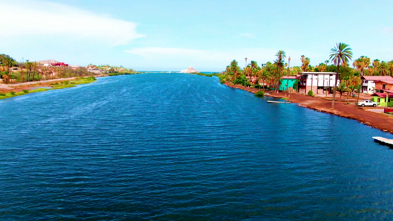 Opening aerial frame over the Mulege waterway in Baja California Sur, with blue rippled water in front, shoreline homes and palms to the right, low hills behind, and no visible people.