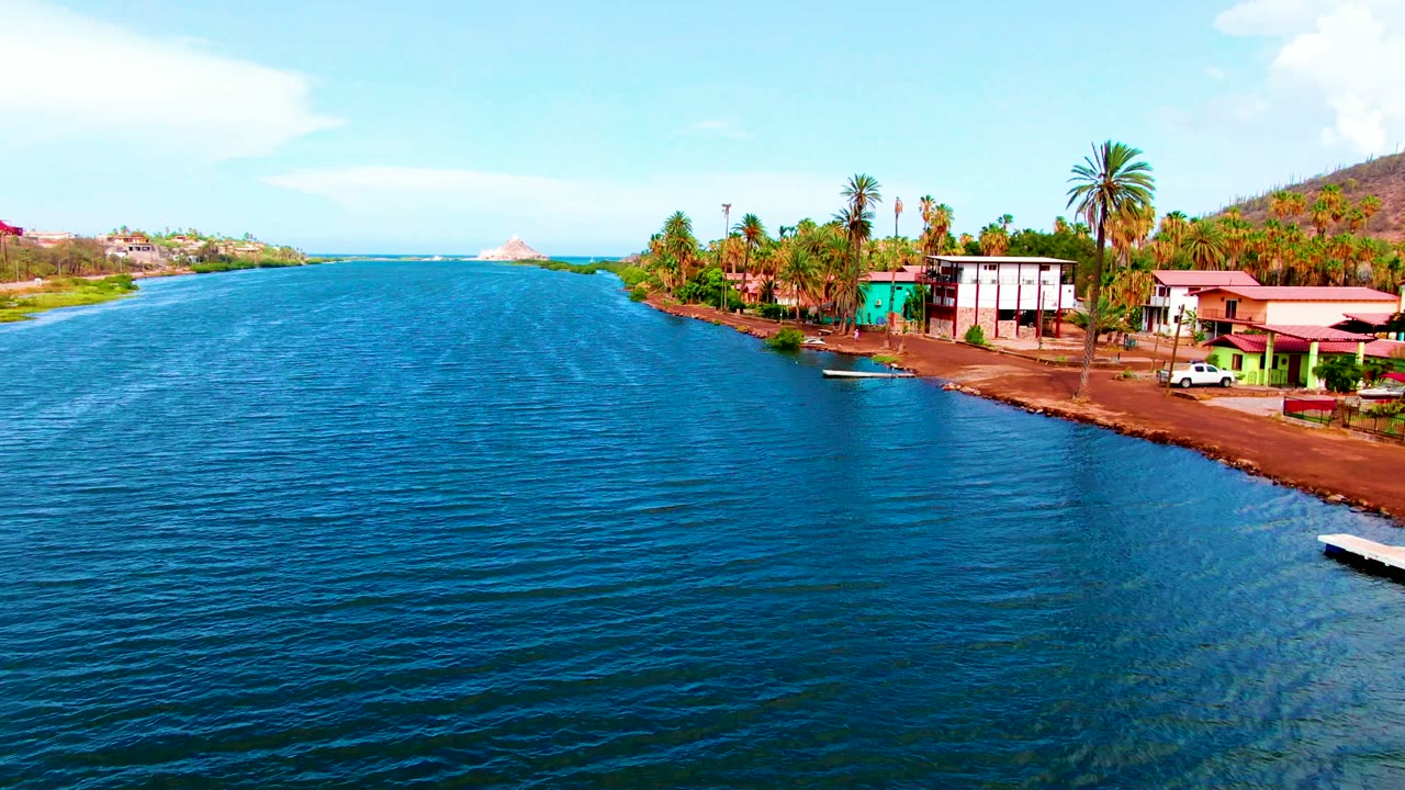 Aerial view along the Mulege shoreline with rippled blue water dominating the foreground, a green building and white waterfront structure near palms, a parked white vehicle, hills, and no visible people.
