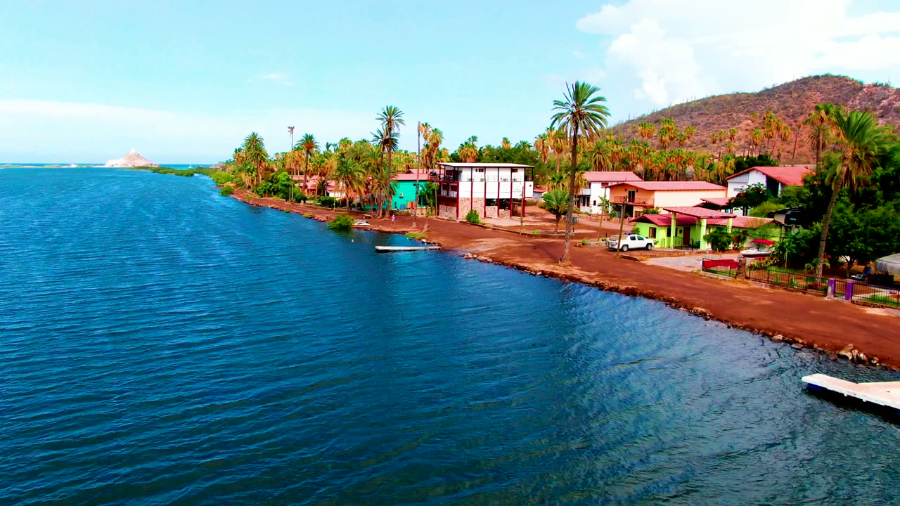 Mulege waterfront frame with the shoreline road running beside blue water, a white multi-story building, red-roofed homes, tall palms, a brown hillside, and no visible people.