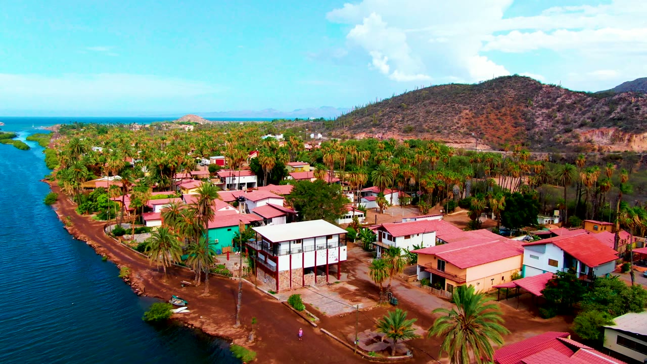 Opening aerial frame over Mulege, Baja California Sur, with blue water at left, palm trees and red-roof homes in the center, dry hills behind, and a tiny distant person on the road.
