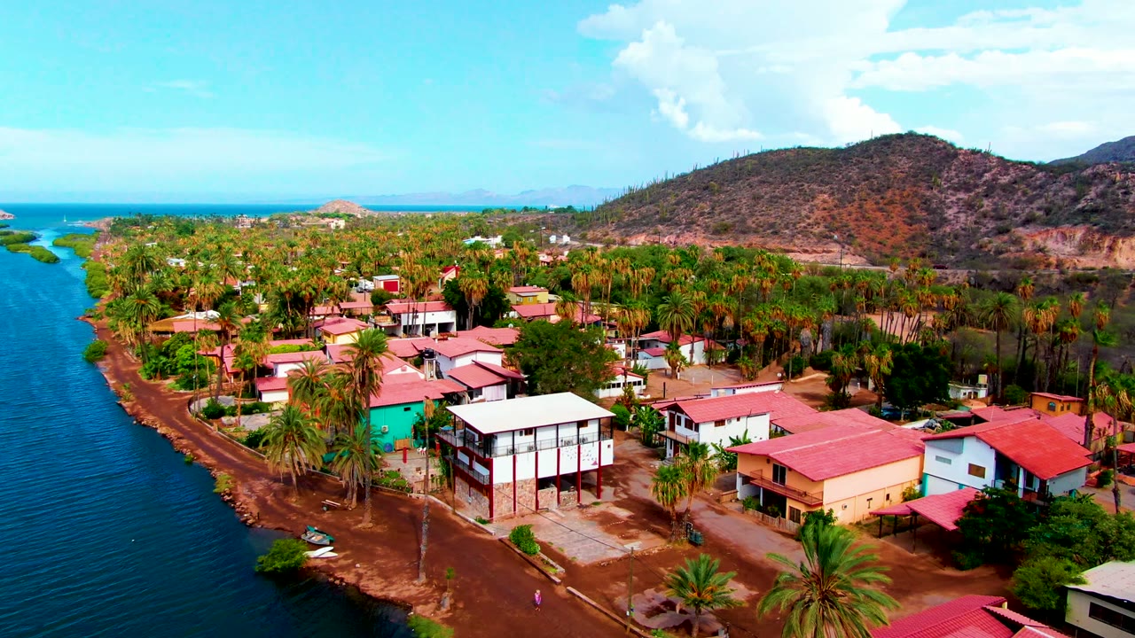 At 0:02, the Mulege village remains framed from above with the blue channel on the left, clustered homes below, palms across the center, and low dry hills beyond.