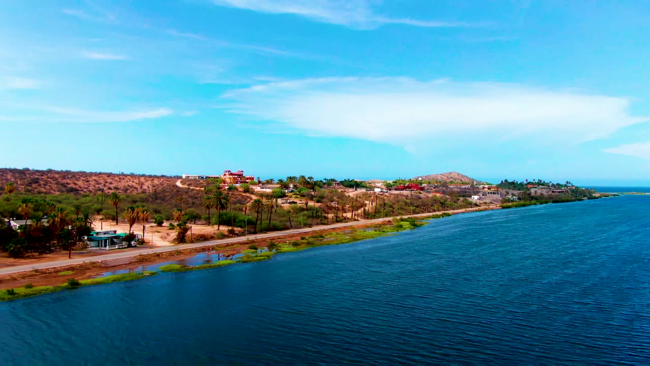 Opening aerial frame in Mulege, Baja California Sur, with blue rippled water in front, a road along the shore, dry hills, scattered palms, low buildings, and no visible people.