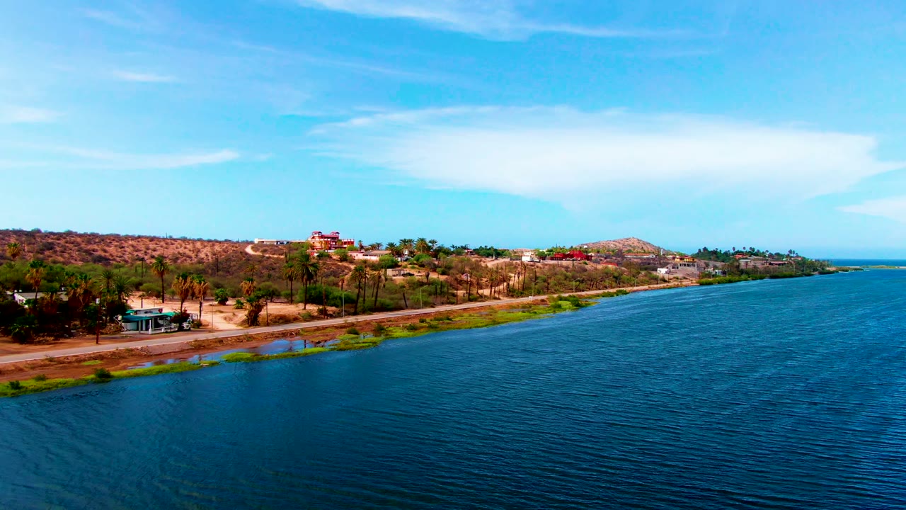 Mulege aerial frame with rippled water filling the right foreground, a narrow road on the shore, dry hillside terrain, palms, low buildings, and no visible people.