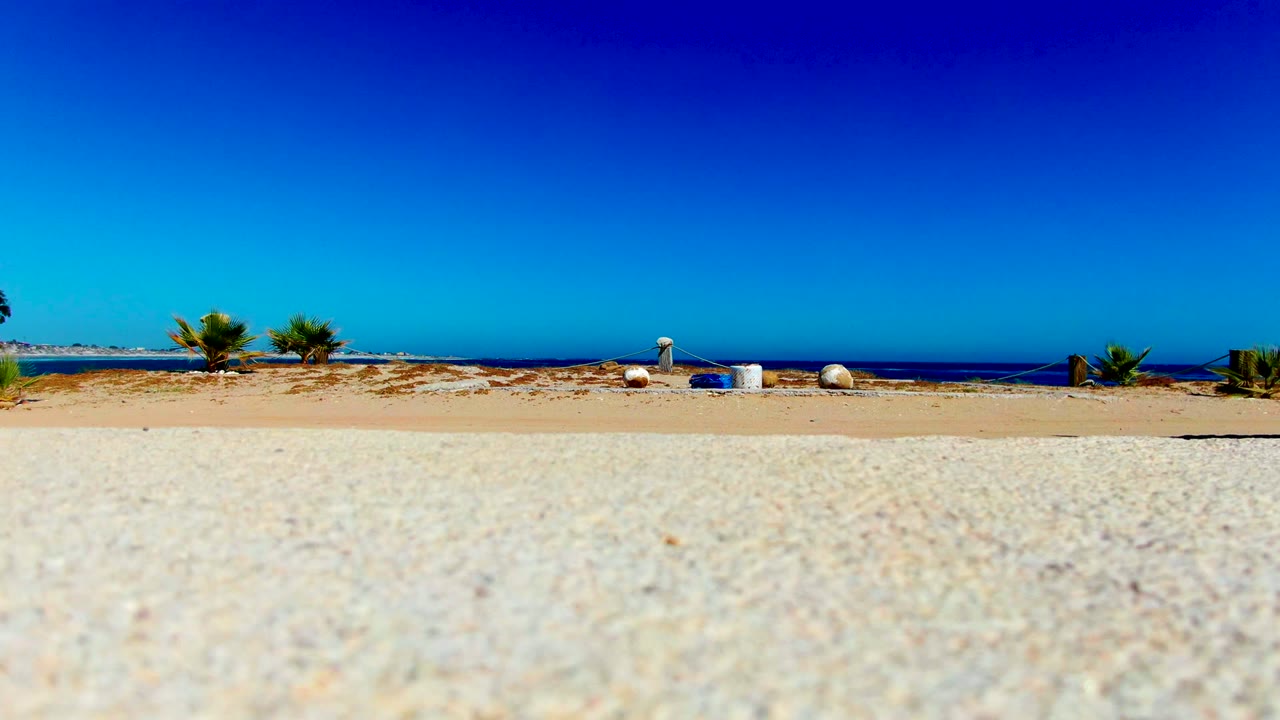 The beach remains dominant in La Bocana, with textured pale sand in front, two small palms and beach objects near the horizon, blue ocean beyond, and no visible people.