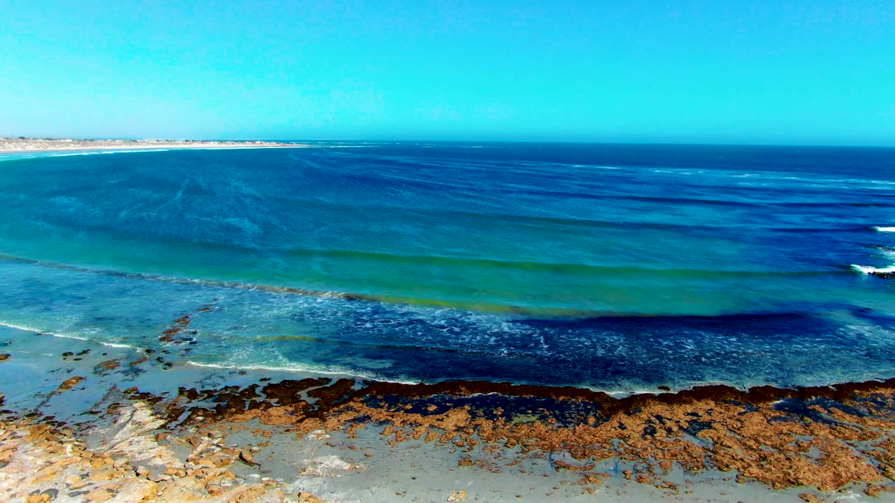 Aerial view in La Bocana, Baja California Sur with turquoise water, dark seaweed patches, white foam near the rocky shore, pale sky, and no visible people.
