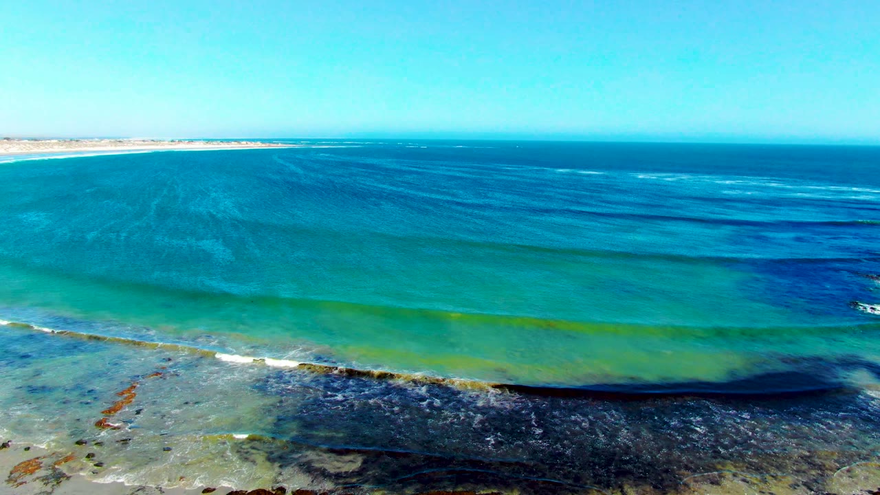 Aerial coastal frame in La Bocana, Baja California Sur with a level ocean horizon, turquoise shallows, white foam over dark nearshore seaweed, and no visible people.