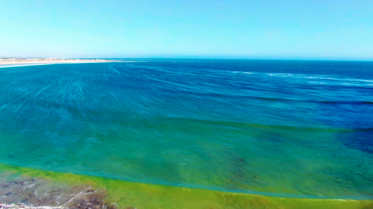 Wide aerial view of turquoise and deep blue water near La Bocana, Baja California Sur, with a thin sandy shoreline at the horizon and no visible people.