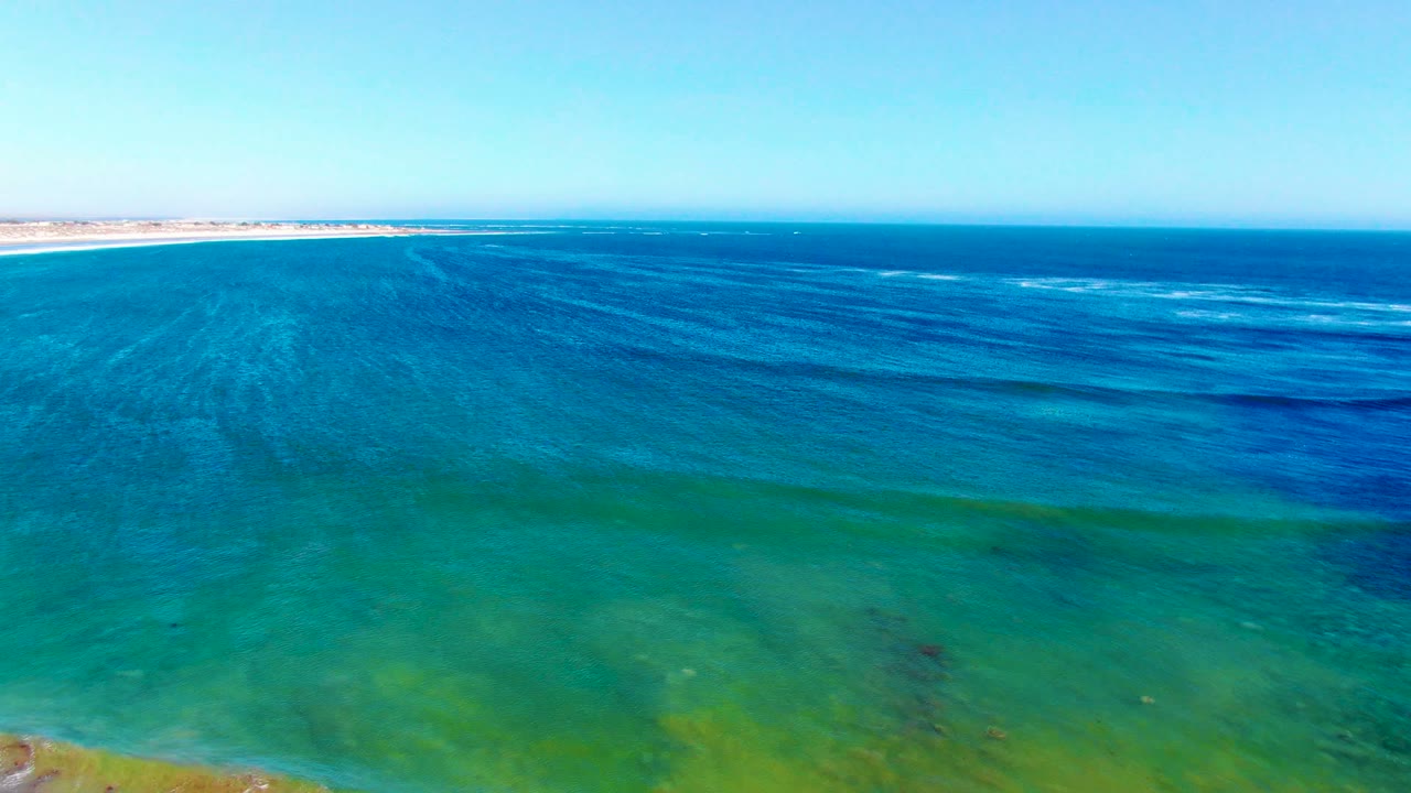 Aerial frame over clear blue-green ocean at La Bocana, with shallow green water in the foreground, darker blue bands offshore, sandy shore at upper left, and no visible people.