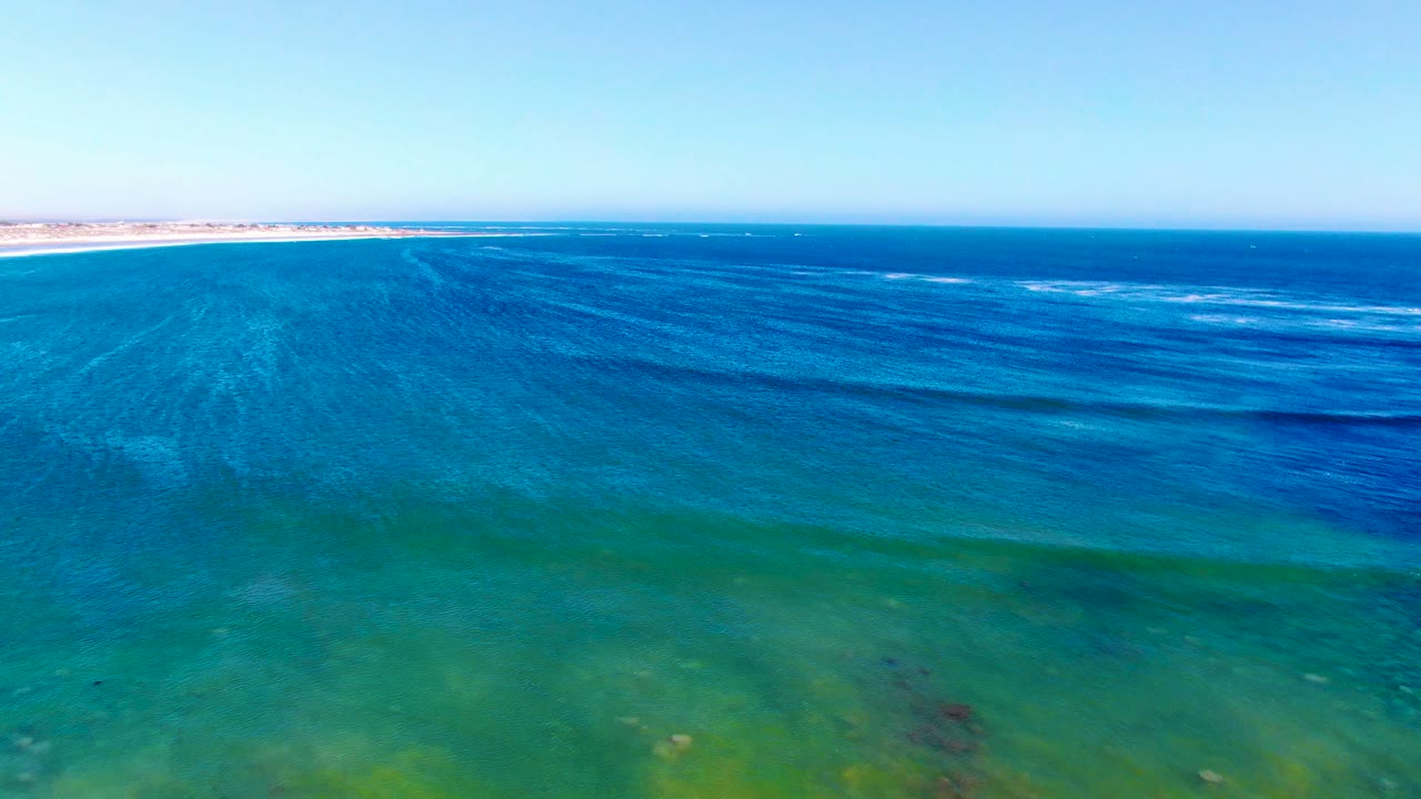 Wide aerial composition over La Bocana coastal water, showing turquoise shallows below, darker ocean bands across the middle, small white wave lines near the horizon, and no visible people.