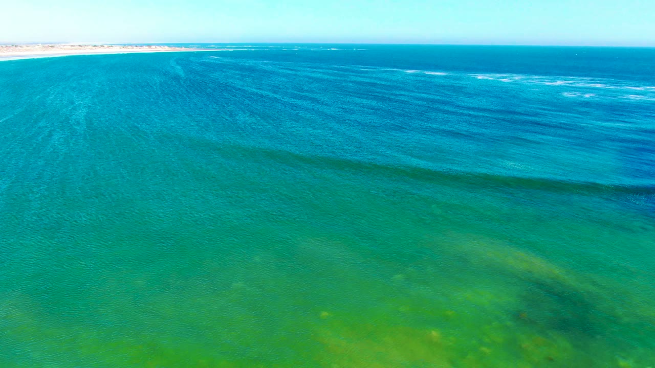Aerial view over clear turquoise water at La Bocana with a thin sandy shoreline near the top edge, green seabed tones below, and no visible people.