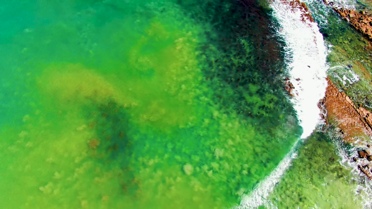 Aerial view of green water and white surf breaking along rust-colored rocks at La Bocana, Baja California Sur, Mexico, with no visible people.