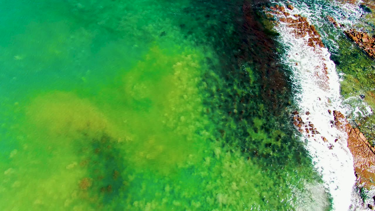 Aerial view at La Bocana showing white surf spread over orange-brown rocks and dark reef patches beside green shallow water, with no visible people.