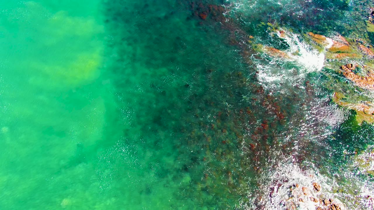 Overhead view of turquoise water and white foam breaking over dark rocky shoreline at La Bocana, Baja California Sur, with no visible people.