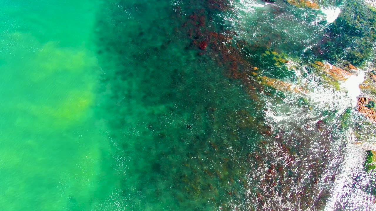 Aerial view at La Bocana showing a bright patch of white foam curling across rocks near the upper right, surrounded by clear turquoise and darker reef-like water, with no visible people.