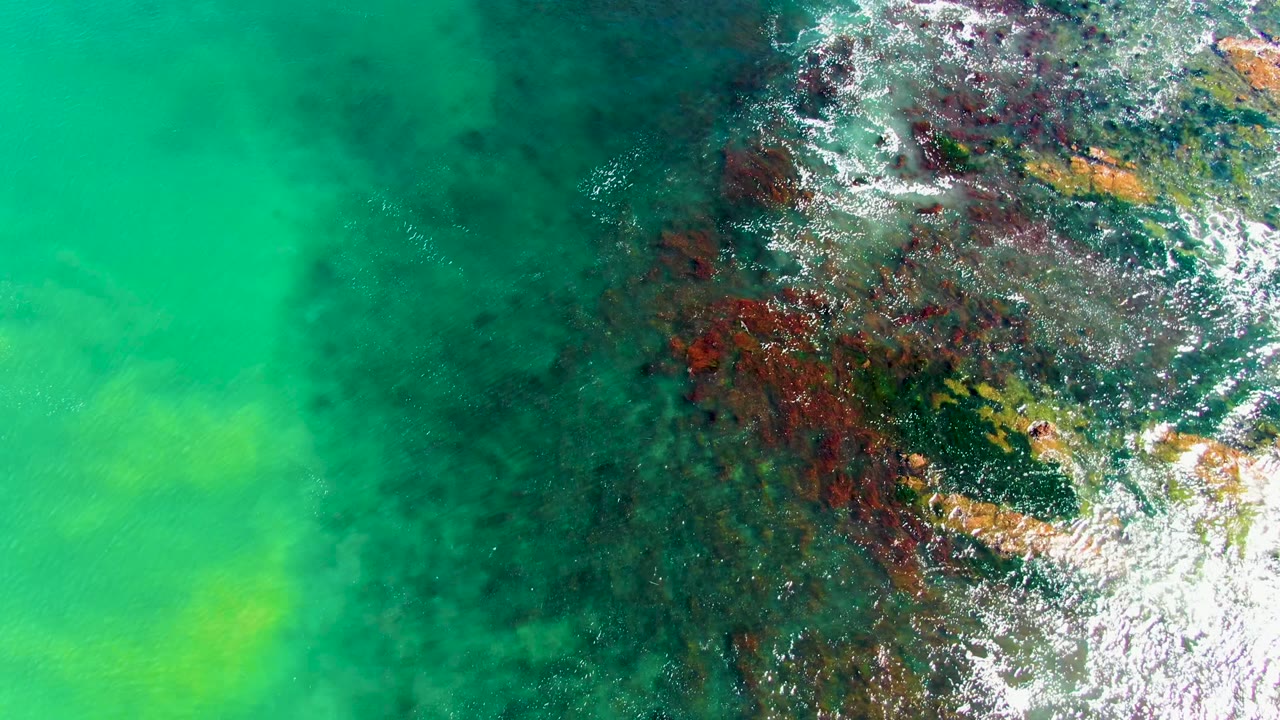 Top-down aerial frame at La Bocana showing turquoise shallow water, dark submerged rocks, white foam along the right edge, and no visible people.