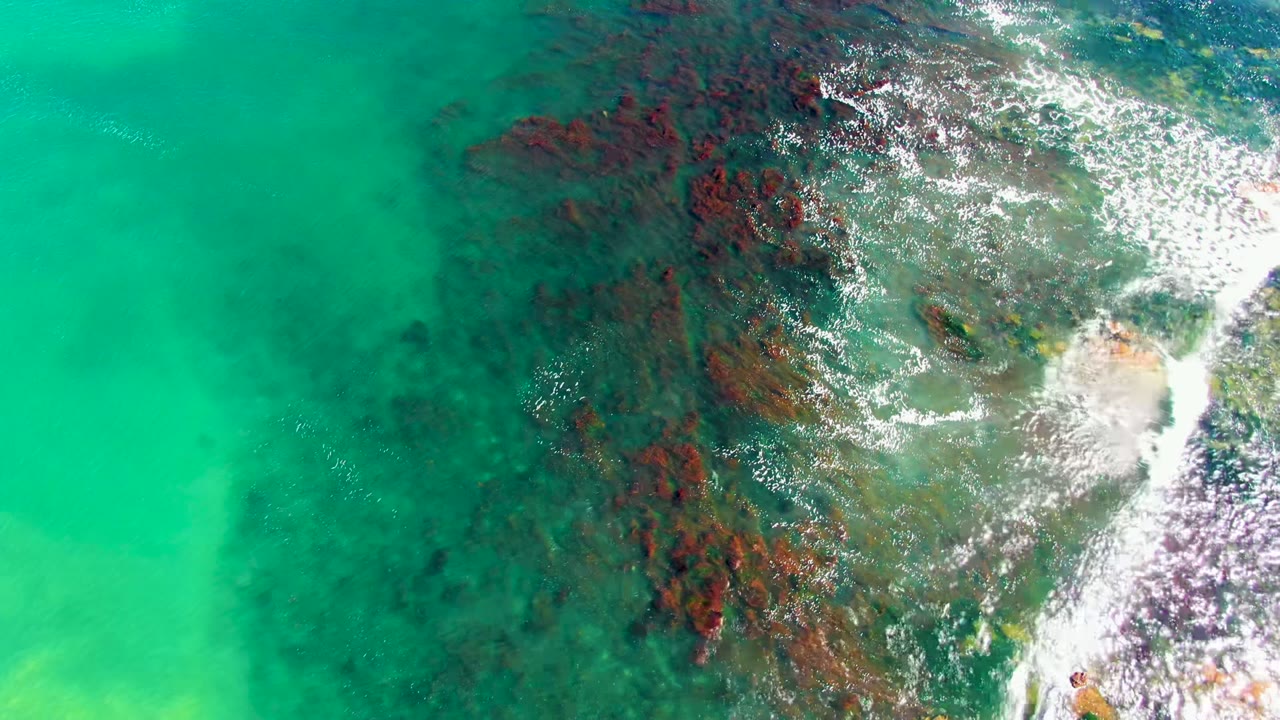 Top-down view of turquoise ocean water in La Bocana, Baja California Sur, with dark red-brown seabed rocks and white foam entering from the right, no visible people.