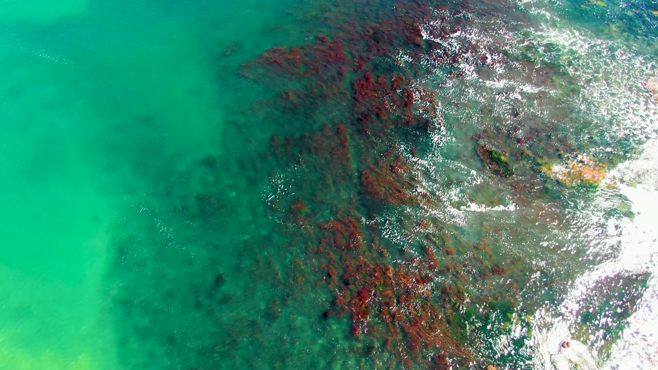 Top-down La Bocana ocean frame with bright turquoise water on the left, darker rocky seabed through the center, and white foam clustered along the right edge, no visible people.