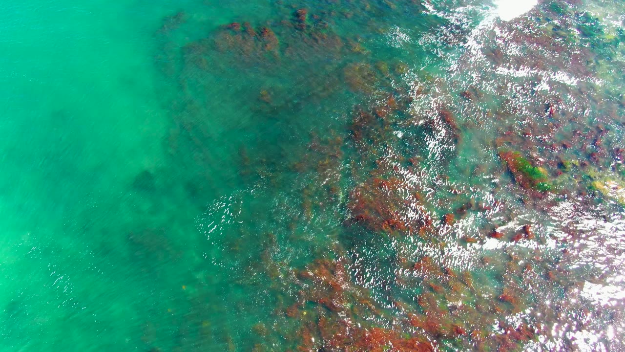 Overhead view of turquoise shallow water in La Bocana, Baja California Sur, with dark submerged rocky formations clustered toward the right and no visible people.