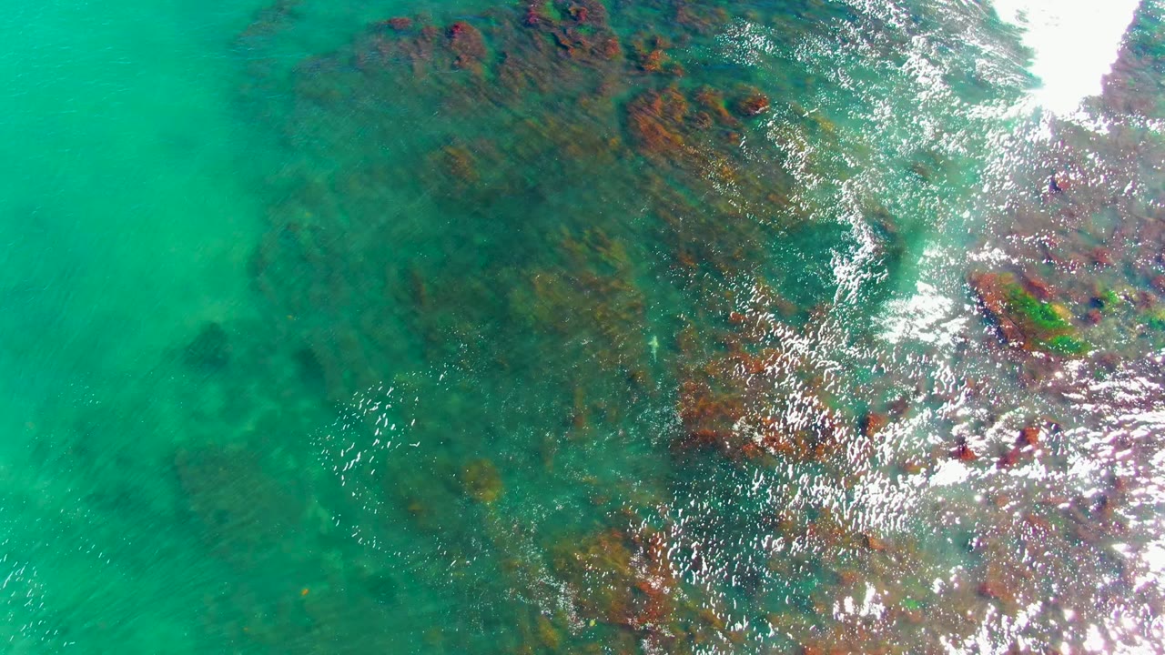 An overhead La Bocana water frame shows darker submerged formations beneath turquoise water, with a larger bright glare area along the upper right and no visible people.
