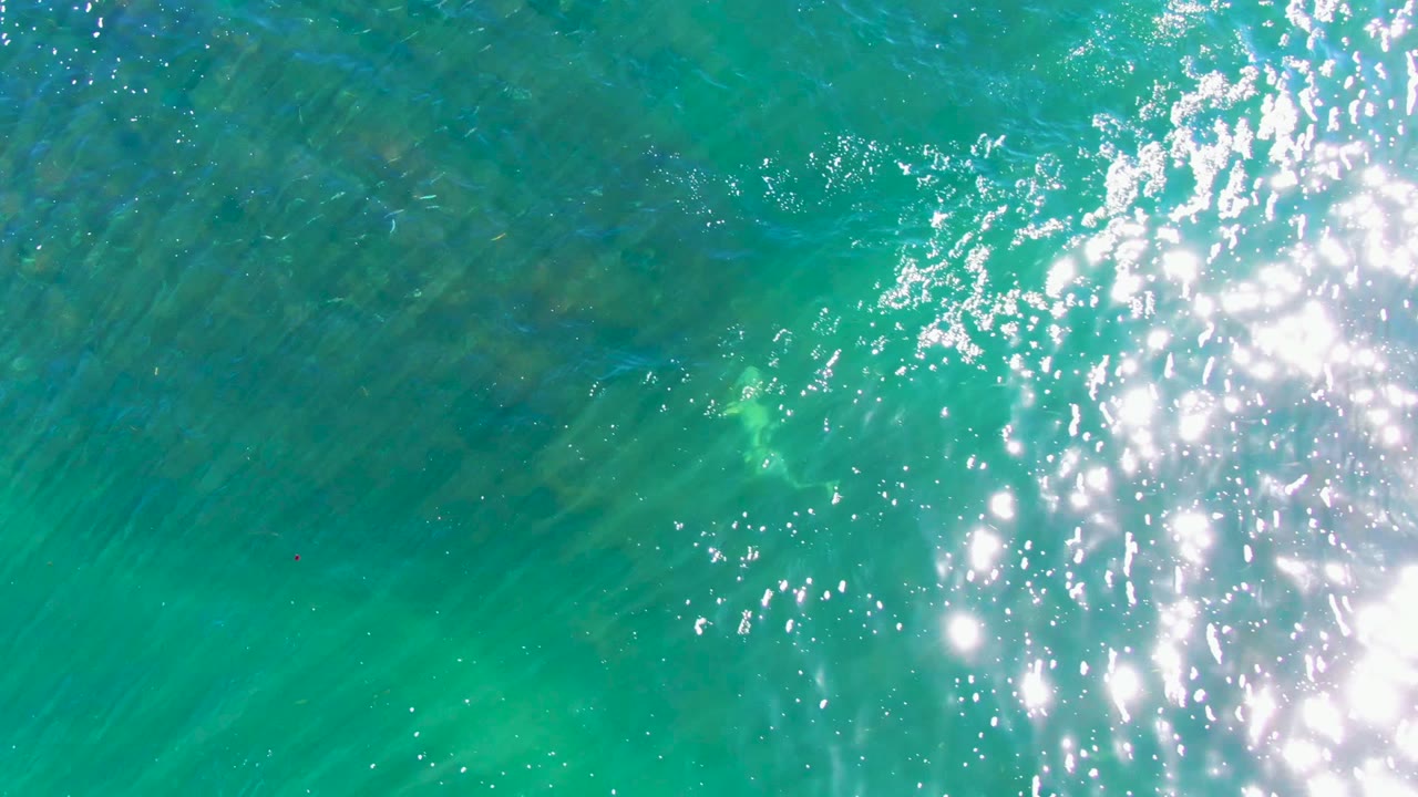 Bright sun glare flashes along the right edge as a faint ray shape sits below the clear turquoise water in La Bocana, Baja California Sur, with no visible people.