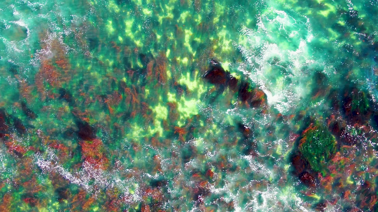 Turquoise shallow water with bright seabed patches and foam over rocks.