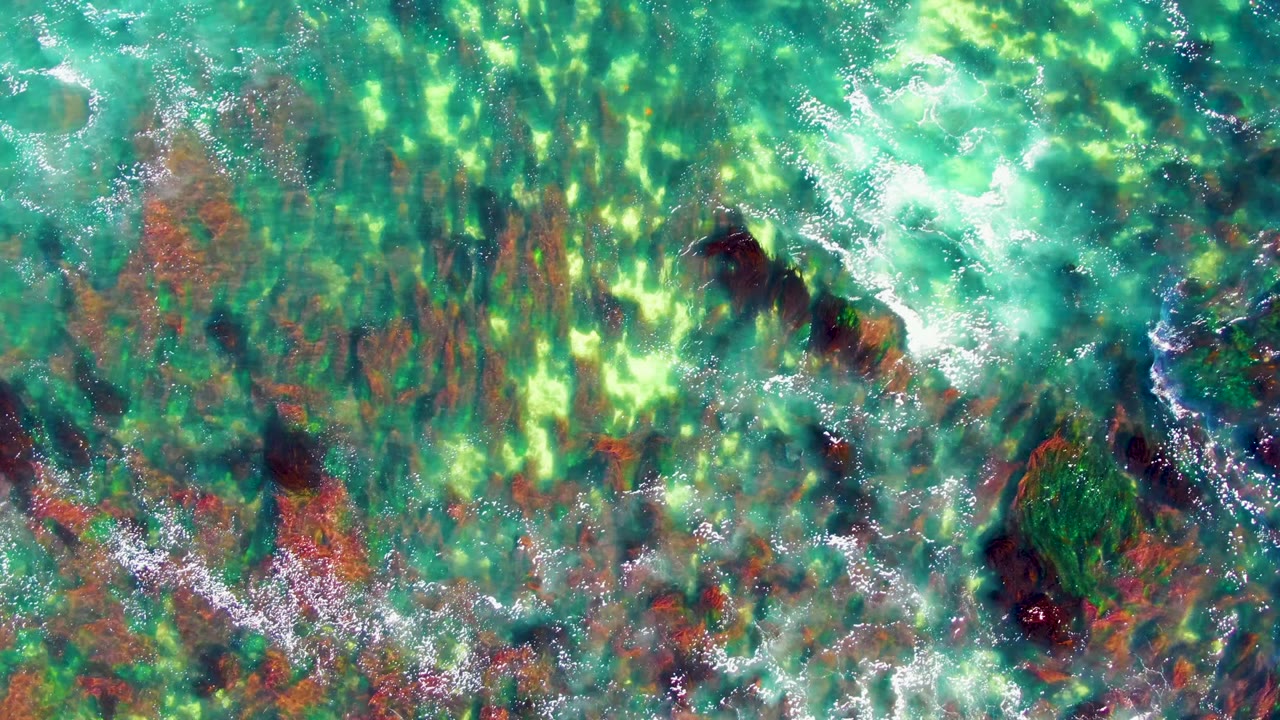 Top-down La Bocana frame with clear green-blue water, clustered wave foam, reddish seabed areas, dark submerged rocks, and no visible people.
