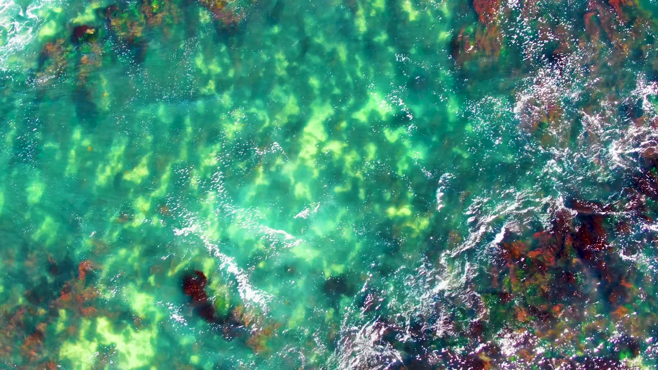 Top-down La Bocana ocean view with bright turquoise water, darker underwater rock clusters, white foam along the right edge, and no visible people.