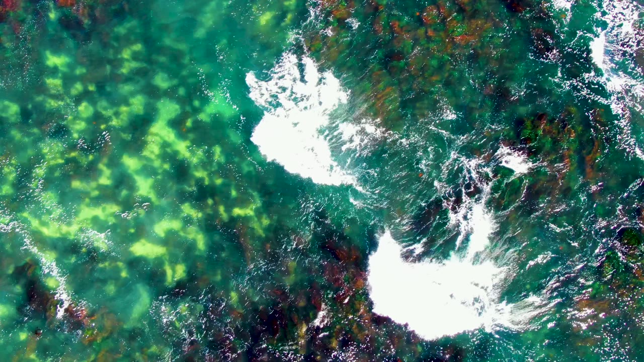 Overhead view in La Bocana, Baja California Sur showing turquoise water, dark submerged rocks, and white foam spreading across the lower center, with no visible people.