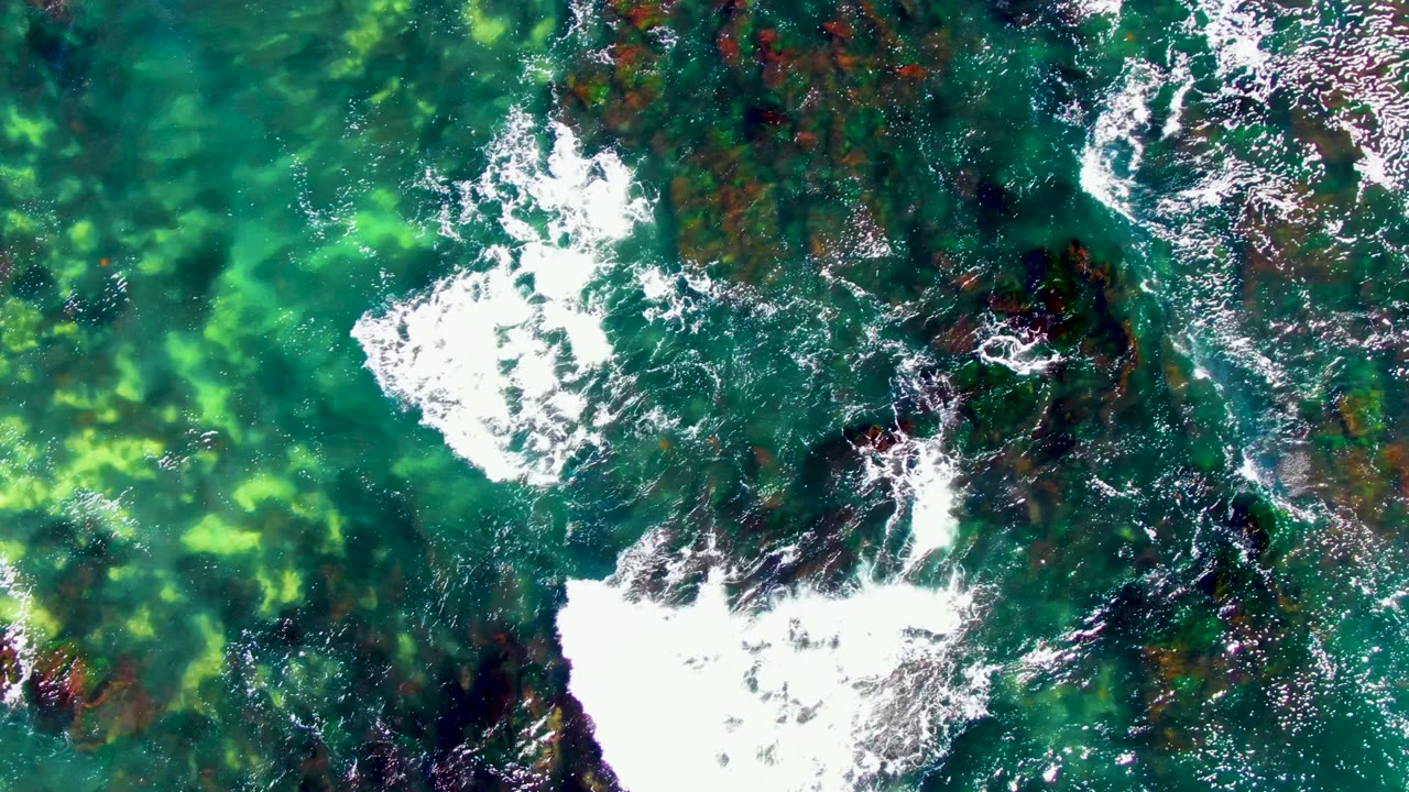 Overhead La Bocana frame with bright white surf breaking across dark rocks while green shallow water shows lighter seabed patches, with no visible people.