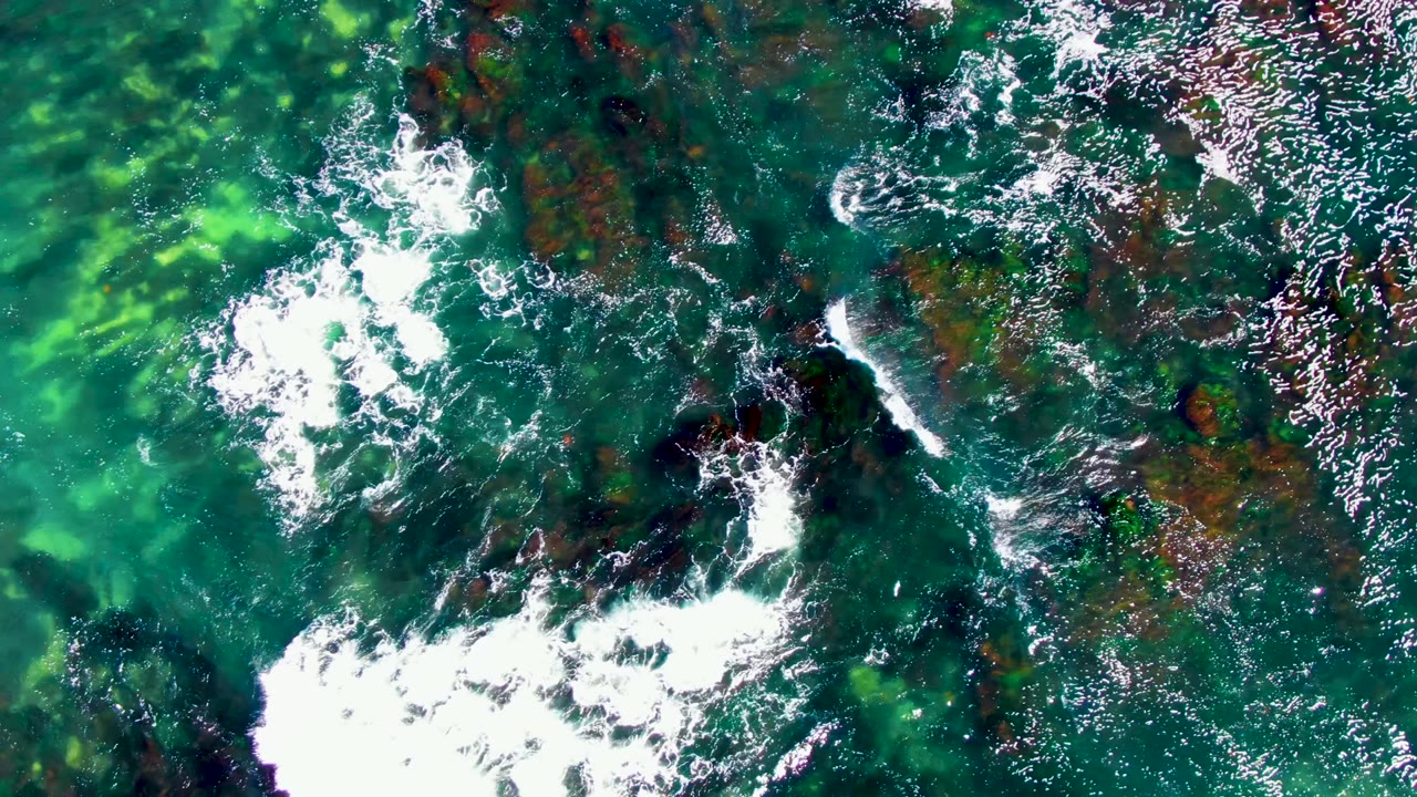 A straight-down view in La Bocana shows a white wave line crossing turquoise water beside dark underwater rock clusters, with no visible people.