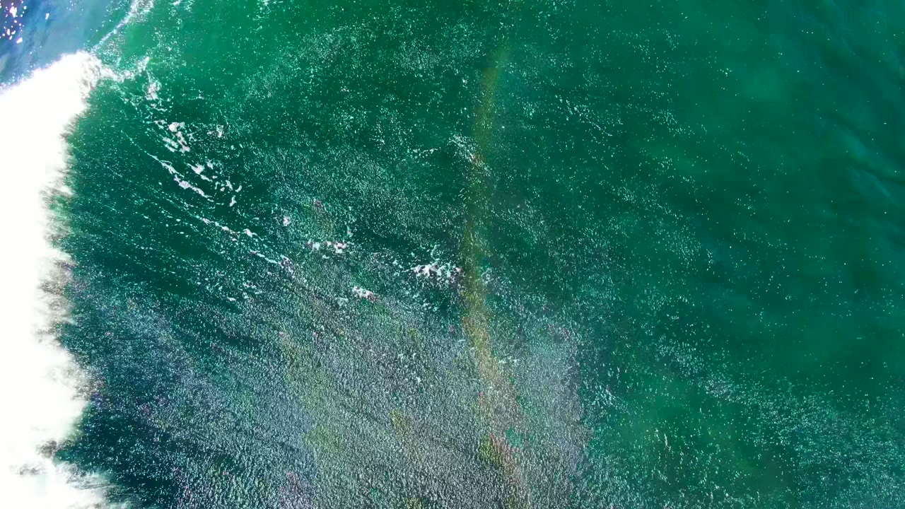 Overhead view at La Bocana with dense white foam along the left edge, darker teal water, a faint rainbow in spray or glare, and no visible people.