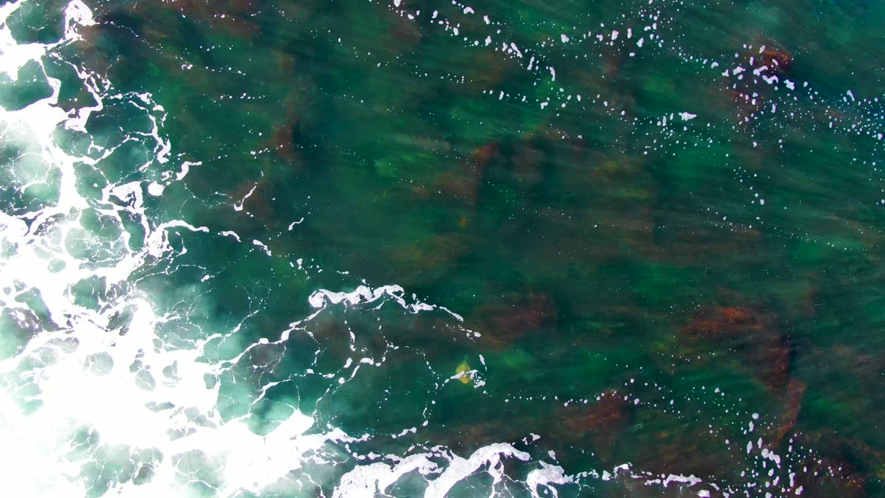 Overhead view of green-blue shallow water at La Bocana, Baja California Sur, with white foam entering from the left and darker submerged rocks visible, no visible people.