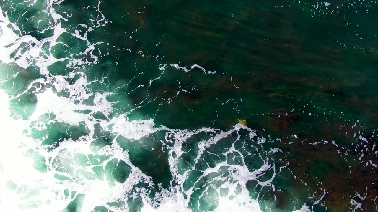 White surf foam spreads diagonally through dark green water at La Bocana, with mottled seabed color visible beneath the surface and no visible people.