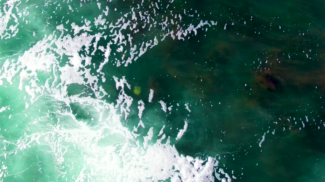 Overhead view of turquoise and dark green ocean water at La Bocana, Baja California Sur, with white foam sweeping across the frame, a small yellow object near the left edge, and no visible people.