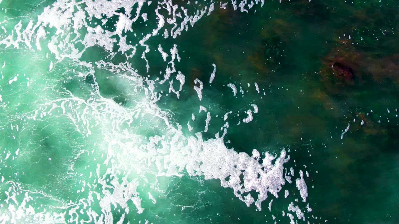 Aerial overhead view of La Bocana water with white foam clustered through the center and lower right over shifting turquoise and dark green sea, with no visible people.
