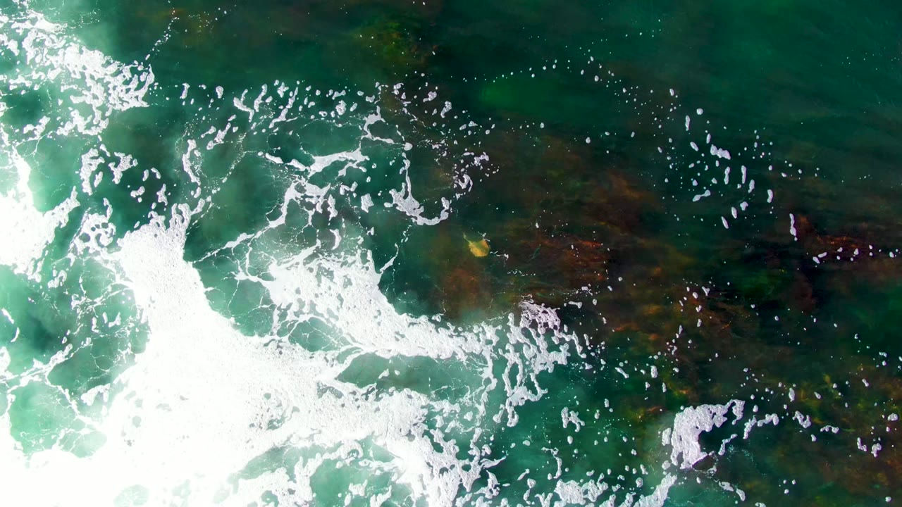 Overhead view in La Bocana, Baja California Sur, showing white surf foam crossing green water with dark rocky seabed patches below and no visible people.