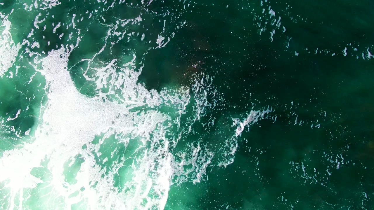 Top-down La Bocana ocean view with white foam clustered across the center, green water around it, faint submerged rocks, and no visible people.