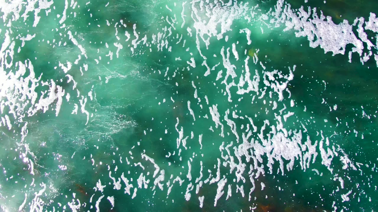 Aerial view over La Bocana water with turquoise color, white foam clusters, and darker submerged seabed patches, with no visible people.