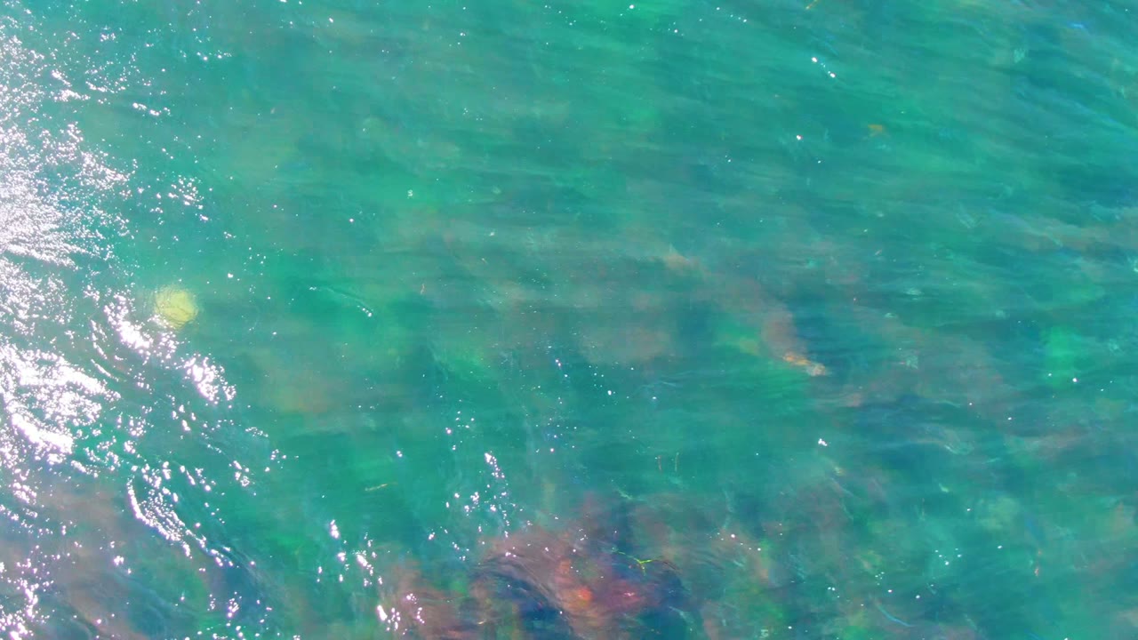Top-down view of turquoise shallow water in La Bocana with faint submerged rocks near the lower edge, bright glare at left, and no visible people.