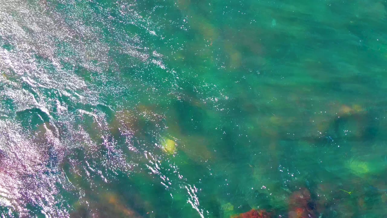Turquoise water at La Bocana fills the frame with scattered submerged rocks, a small yellow object near center-left, surface sparkle, and no visible people.