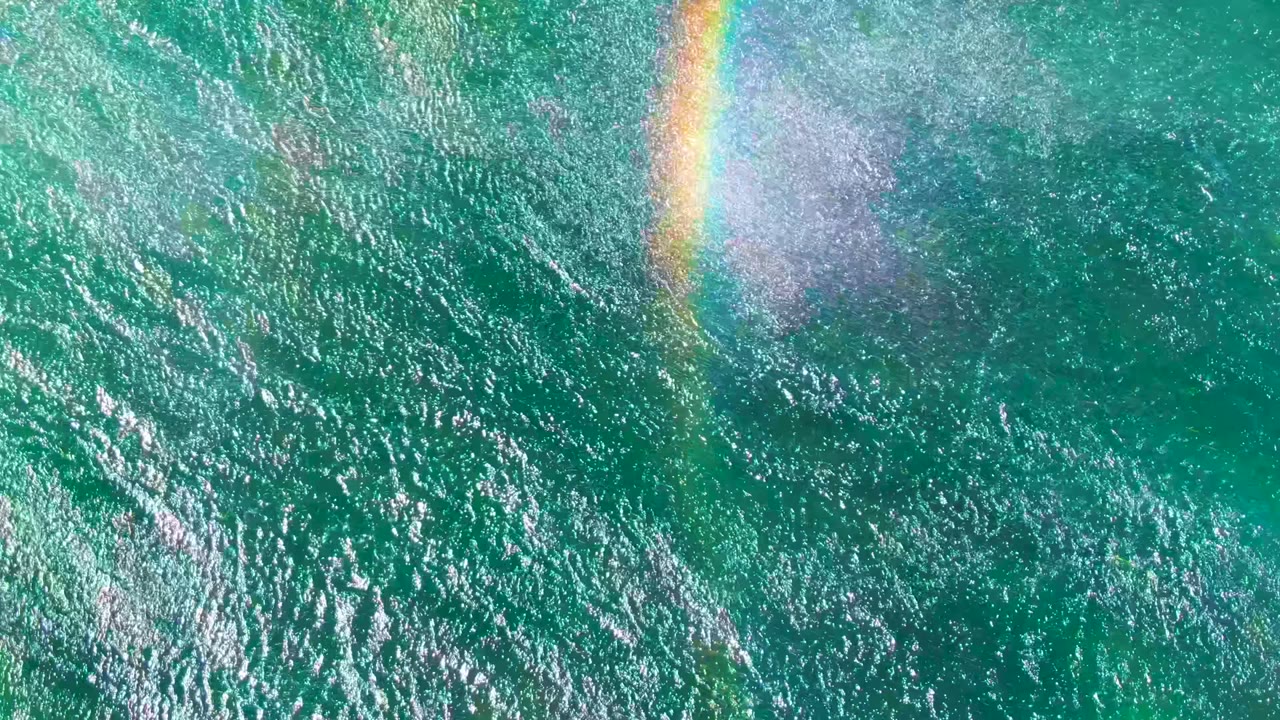 Overhead La Bocana ocean surface with dense sun sparkle, pale spray, and a faint rainbow arc over green-blue water, with no visible people.