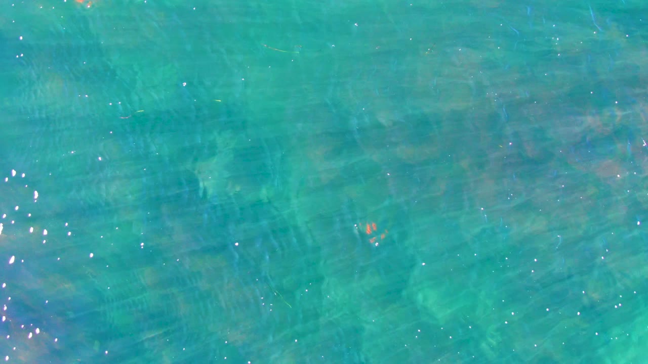 Turquoise coastal water in La Bocana shows subtle wave bands, muted green underwater patches, and a narrow vertical run of bright sun reflections, with no visible people.
