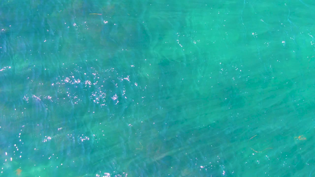 Top-down turquoise ocean water in La Bocana with small ripples, faint underwater streaks, scattered sun reflections, and no visible people.