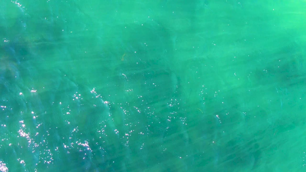Turquoise ocean surface in La Bocana, Baja California Sur, fills the frame with ripples, clustered sun glints, faint fish below the surface, and no visible people.